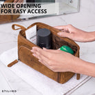 Necessaire em couro na cor tan/marrom escuro, abertura larga com interior visível, mãos organizando produtos, logotipo STILORD