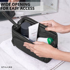 Necessaire em couro preto com abertura ampla, vista interior com mãos e produtos de higiene sobre toalha, gravação STILORD visível